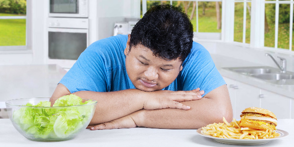 a large asian man sits with his head in his hands as he weighs up whether to eat lettuce or a plate full of chips and a burger, representing obesity in hong kong